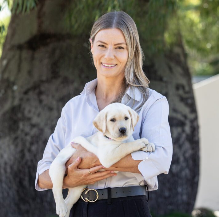 Image shows Victoria smiling at the camera, holding a Labrador puppy.
