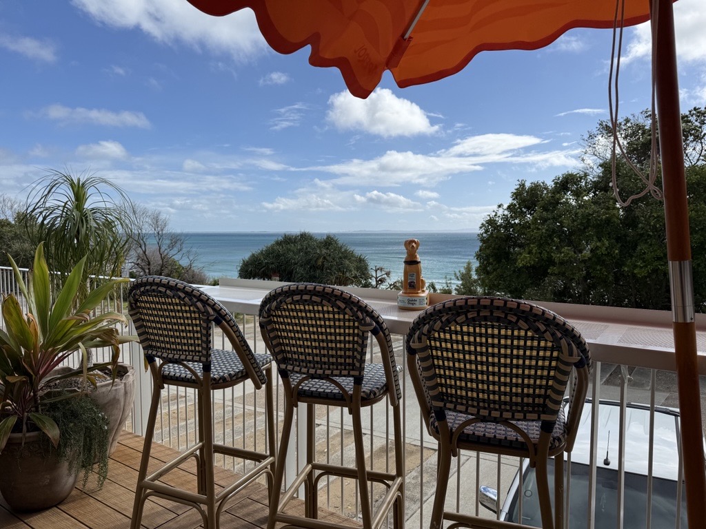 A view from a deck over a white railing looking out at the ocean under a cloudy blue sky. Three woven barstools sit at a high table, and an orange umbrella shade is at the top of the frame.