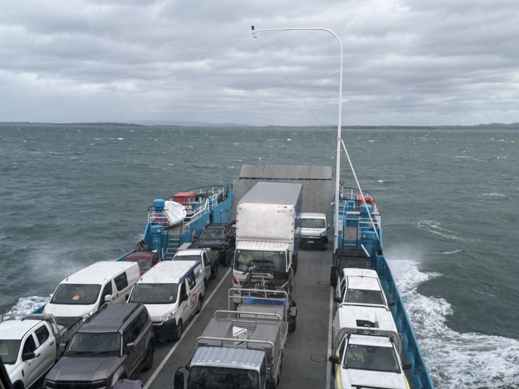 A high-angle view looking down at the packed deck of a vehicle ferry crossing choppy, dark water under a cloudy sky. The deck is filled with various cars, vans, and several white work trucks.