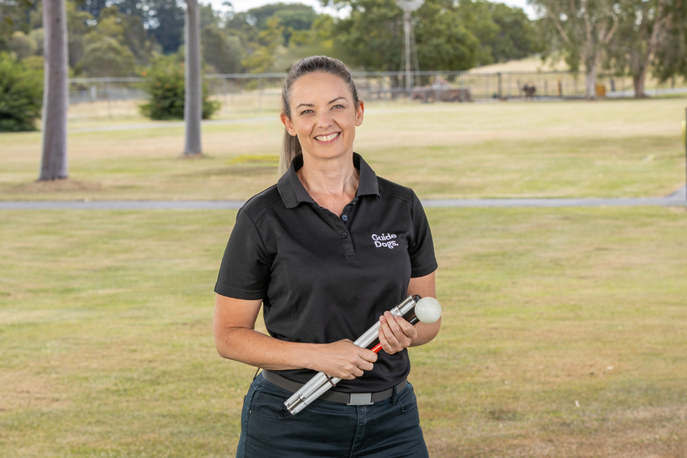 EJ, wearing a black polo shirt with "Guide Dogs" on the chest, stands outdoors holding a folded white cane.