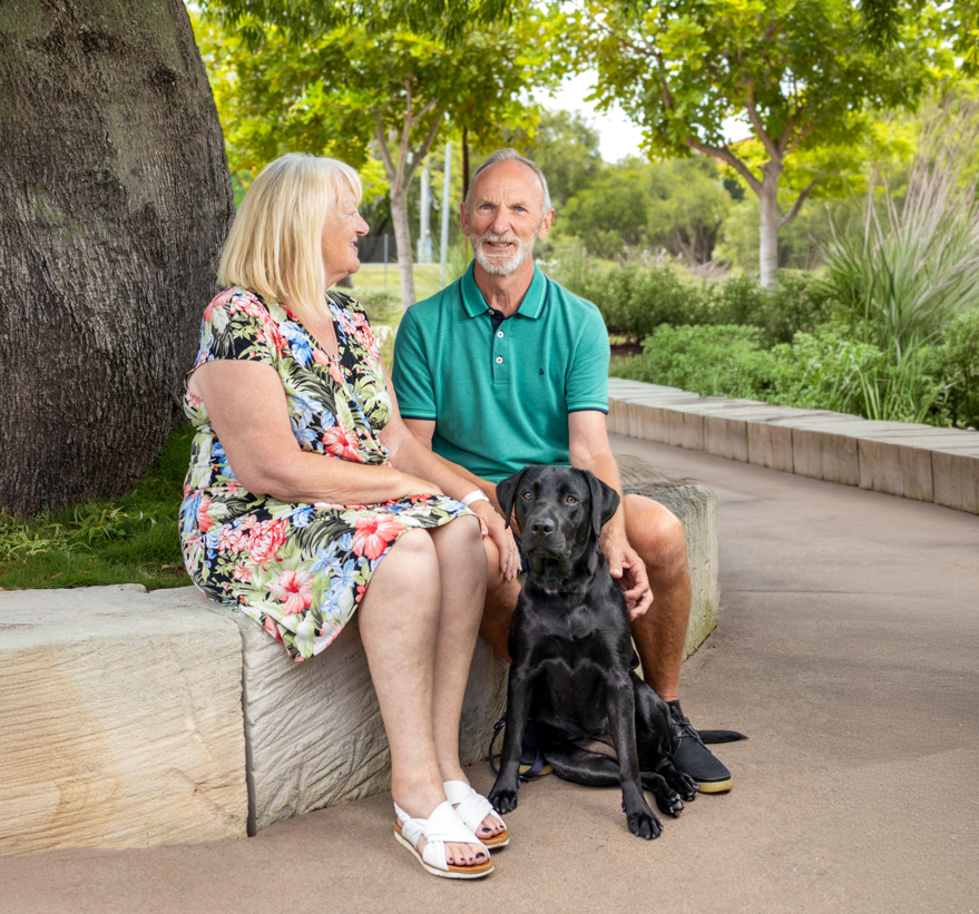 A smiling older man and woman sitting outdoors on a stone bench under a tree, with a black Labrador dog sitting in front of them.