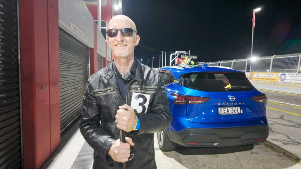 Rhett standing behind a car the the Blind Speed Track Day event holding a white cane.