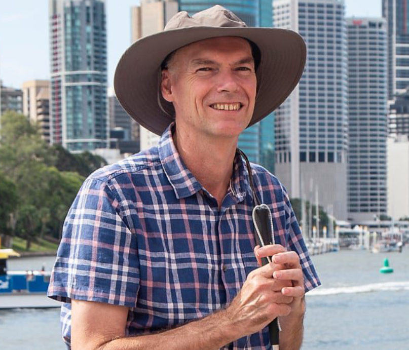A smiling man in a plaid shirt and sun hat holds a white cane against a backdrop of city skyscrapers and a body of water.