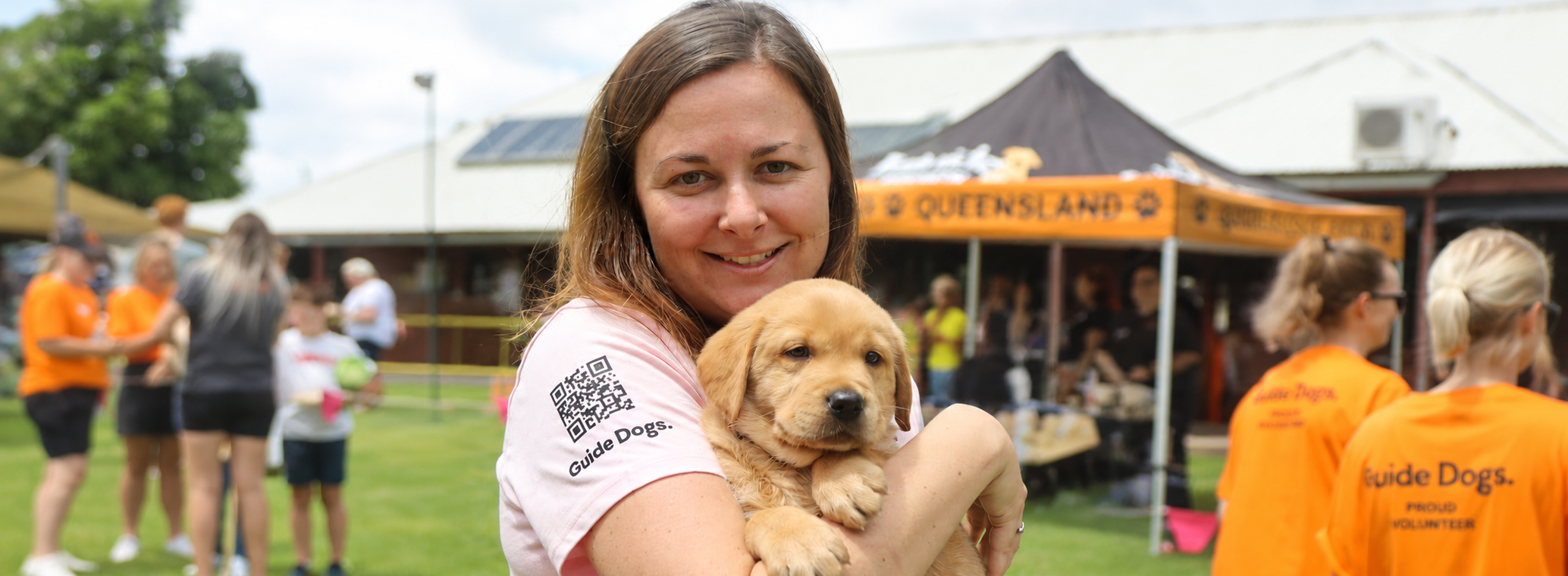 A smiling woman wearing a pink Guide Dogs shirt holds a golden retriever puppy outside, with other people in orange shirts in the background.