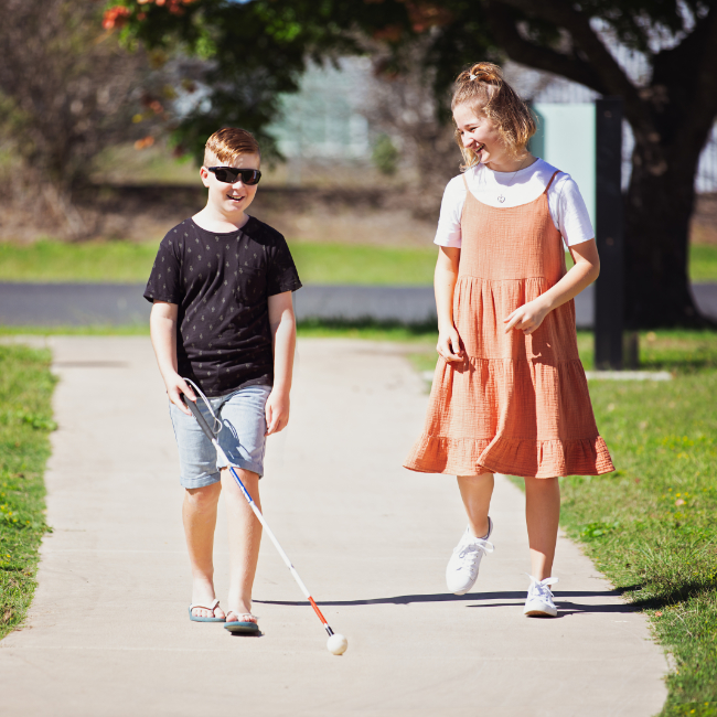 Two children walk together along a footpath, with one child using a white cane.