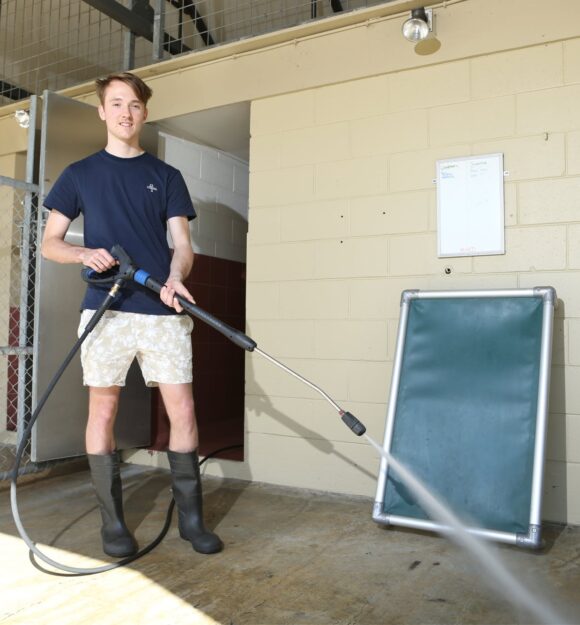 A person standing outside holding a high pressure hose and wearing some gum boots.