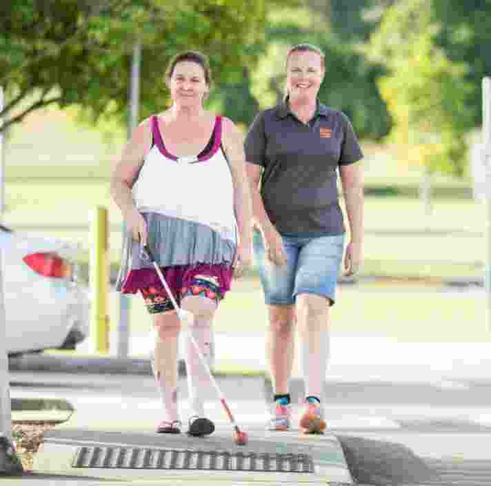 A person crossing the road with their white cane while a Guide Dog's staff member walks beside them. They are both looking at the camera smiling.