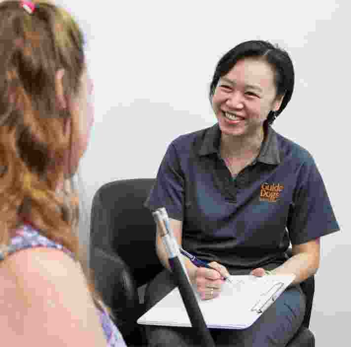 A person sitting with a Guide Dog's staff member. The staff member is smiling while the other person is not facing the camera.