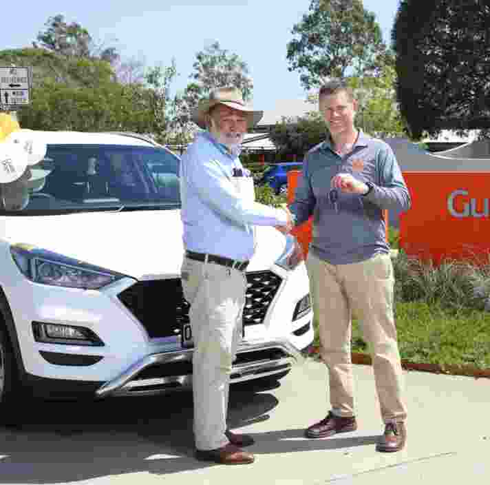 Two people shaking hands in front of a white car. The car has yellow, orange and white balloons stuck to the driver side door. Both people are looking at the camera smiling.