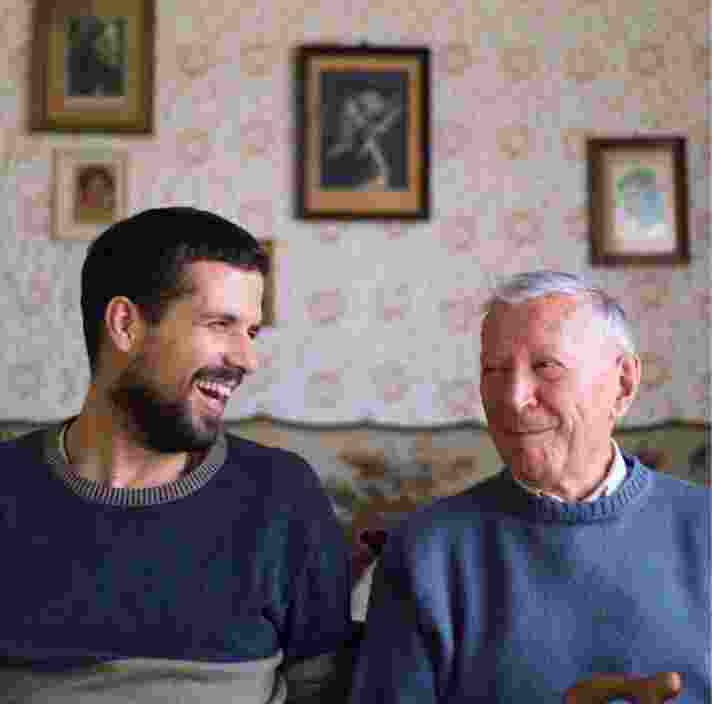 Two people, one adult & one older person, sitting on a couch looking at each other laughing.