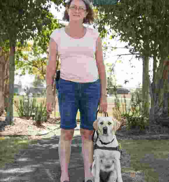 A person standing on a footpath with their yellow Guide Dog in harness. Both the Guide Dog and person are looking at the camera.