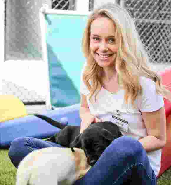 A person sitting outside smiling at the camera while two eight week old puppies, one yellow and one black, play together in their lap.