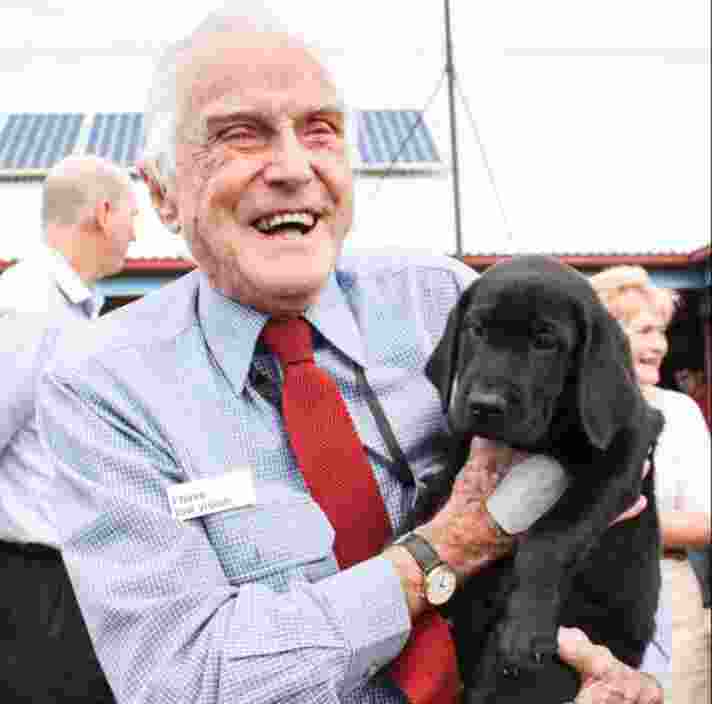 A Guide Dog's staff member holding a black eight week labrador puppy. The staff member is smiling at the camera.