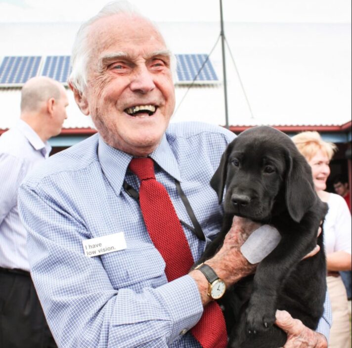 A Guide Dog's staff member holding a black eight week labrador puppy. The staff member is smiling at the camera.