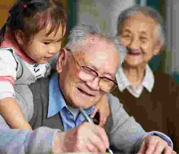 An older man writing while a young girl puts her arms around his shoulders, hugging him. The man is smiling and there is an older woman in the background who is not in focus but is also smiling.