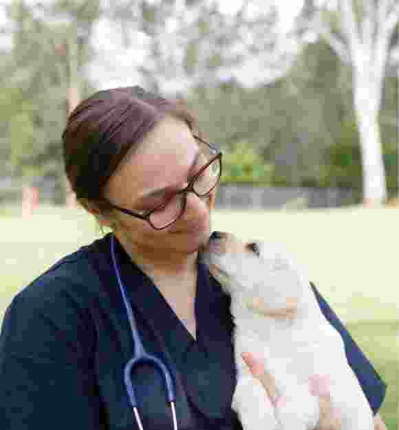 A veterinarian holding an eight week old yellow labrador puppy. The photo is taken outside and the puppy and vet are looking at each other, the veterinarian is smiling at the puppy.