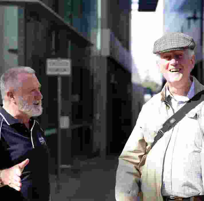 A Guide Dogs staff members is talking to an older adult in the street. The an older adult is looking at the camera with a smile on his face.