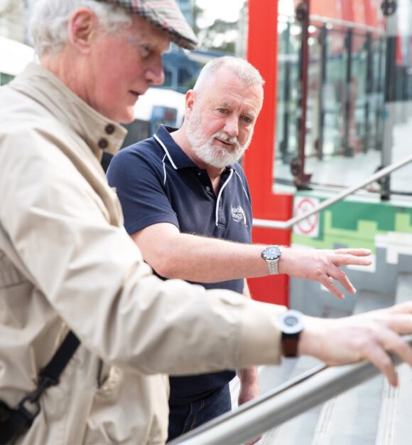 A Guide Dogs staff member assisting an an older adult with navigating a handrail.