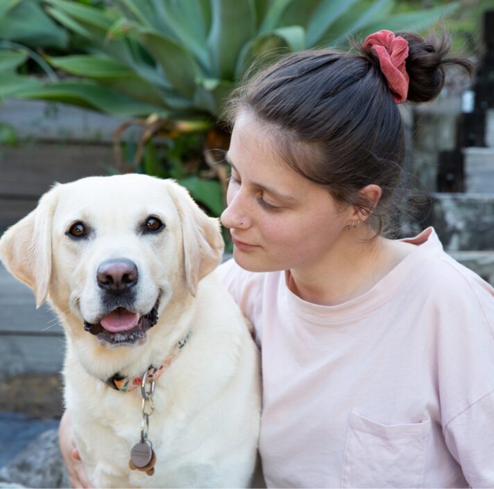 A person seated next to a yellow labrador dog, The person is looking at the dog and the dog is looking at the camera.