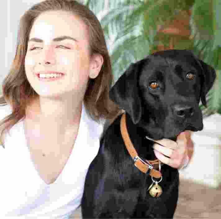A person looking to the left of the camera smiling with their arm around the neck of a black labrador dog.