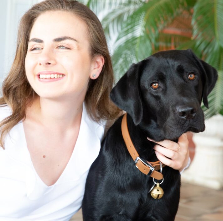 A person looking to the left of the camera smiling with their arm around the neck of a black labrador dog.