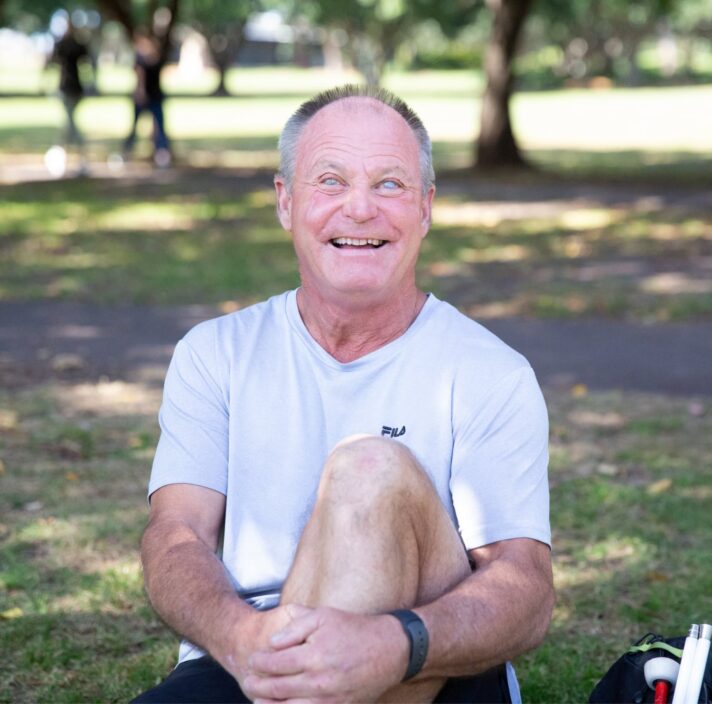 A person sitting outside on the grass with a smile on their face looking at the camera. They have one leg bent with their arm wrapped around their knee and a white cane next to them.
