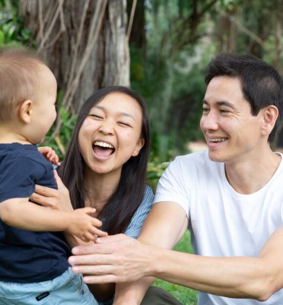 A couple sitting outside watching their baby walk towards them. The mother and father have smiles on their faces.