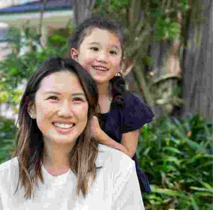 A mother and young child outside smiling at the camera. The young child is standing behind the mother with their hands on her back.