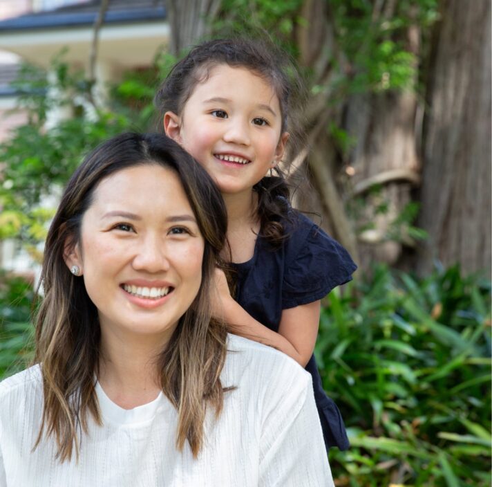 A mother and young child outside smiling at the camera. The young child is standing behind the mother with their hands on her back.