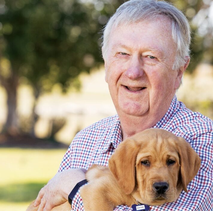 Guide Dogs Queensland Board President, Richard Anderson OAM, standing outside holding an eight week old caramel labrador puppy. Richard is looking at the camera smiling.