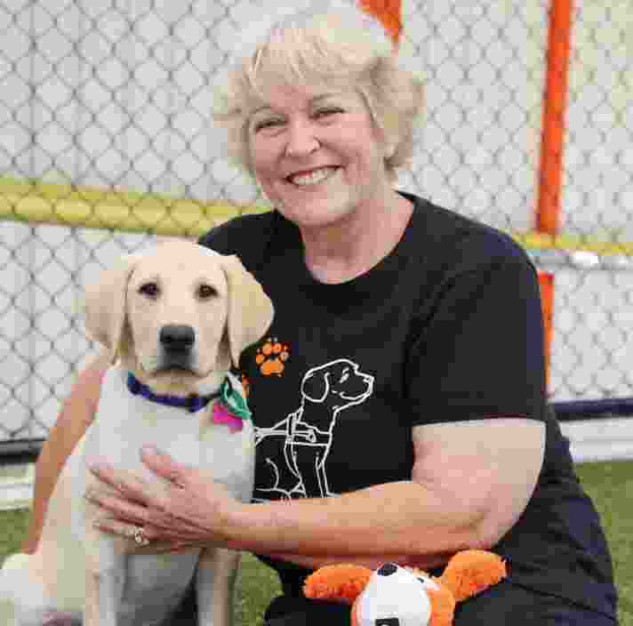 A person sitting on the ground outside next to a 12 week old yellow labrador puppy. The person is smiling at the camera.