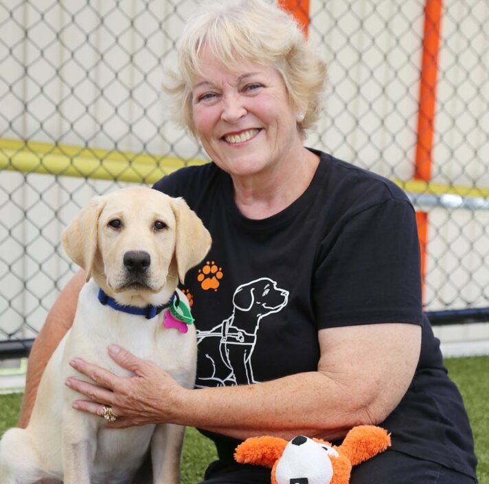 A person sitting on the ground outside next to a 12 week old yellow labrador puppy. The person is smiling at the camera.