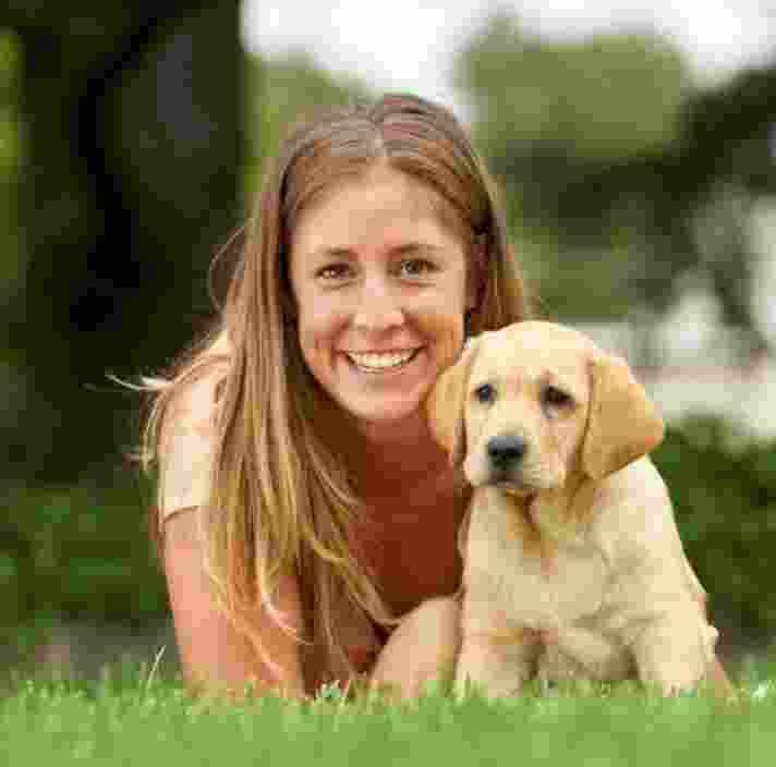 A person laying on some grass with an eight week old yellow labrador puppy. The person is smiling and looking at the camera. The puppy is also looking at the camera.