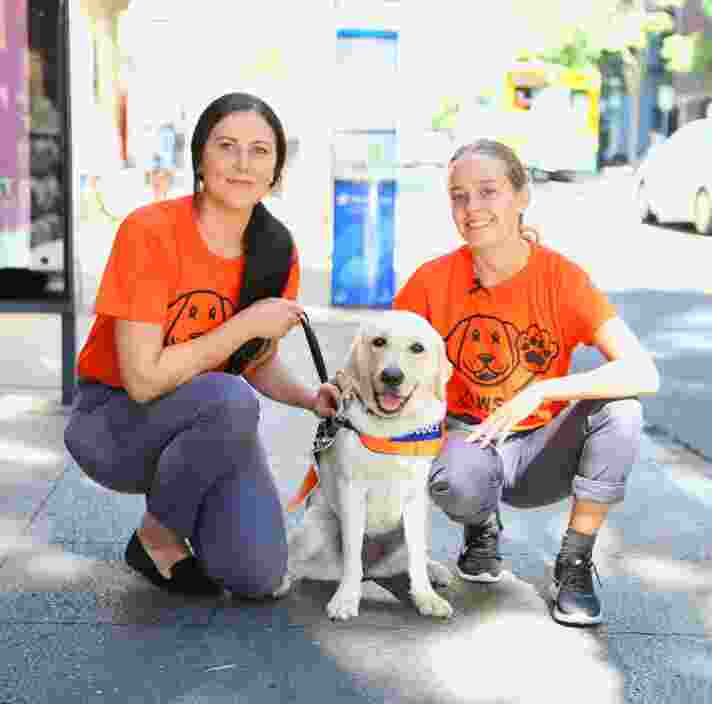 Two people kneeling next to a yellow Guide Dog in training who is wearing a Guide Dog in Training jacket. They are outside on the footpath. The people are smiling while looking at the camera.