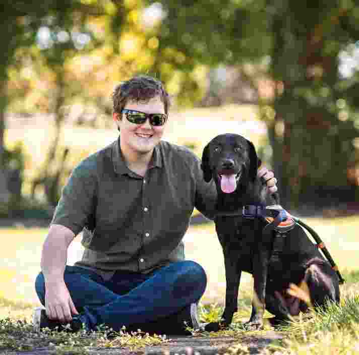 Brent and Guide Dog Jaycee sitting on the grass, looking at the camera, smiling.